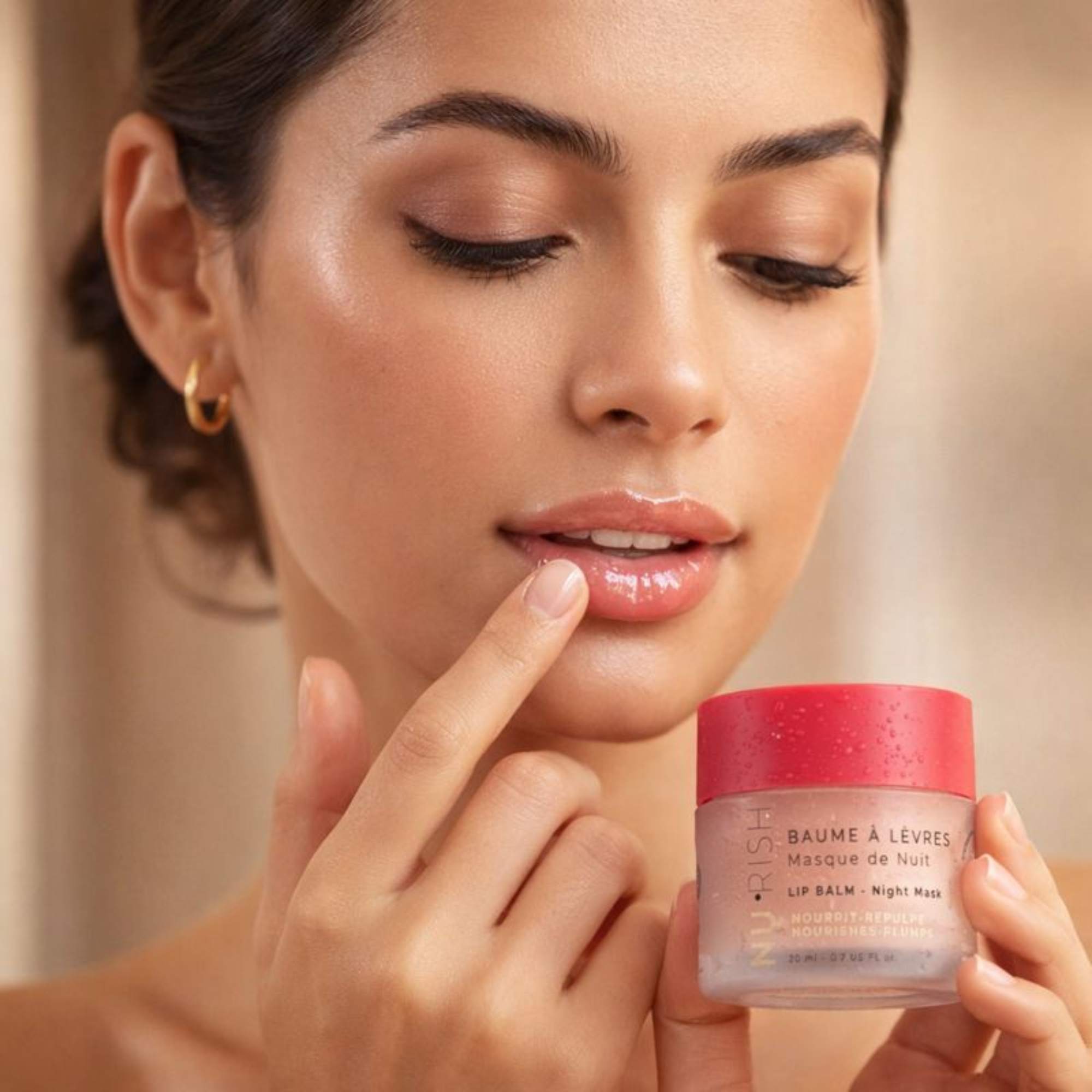 Femme appliquant le baume à lèvres NU•RISH Masque de Nuit avec le doigt, lèvres brillantes et hydratées, tenant le pot au couvercle rose sur fond doux et lumineux.