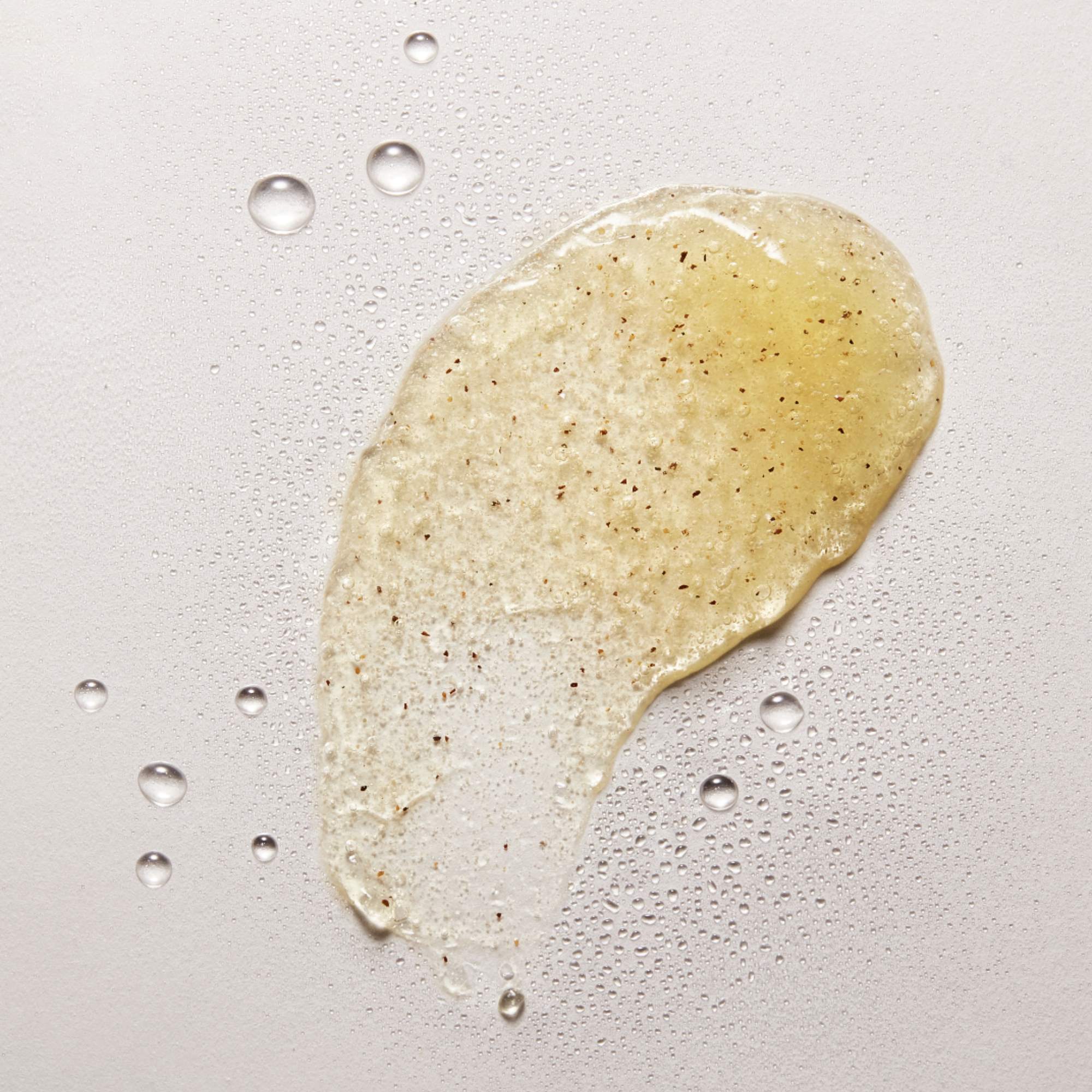 Texture de gel exfoliant NU•RISH appliquée sur fond clair, avec grains exfoliants visibles et gouttelettes d’eau, illustrant une exfoliation douce et hydratante. NU•RISH exfoliating gel texture applied to a light background, with visible exfoliating grains and water droplets, illustrating gentle, moisturizing exfoliation.