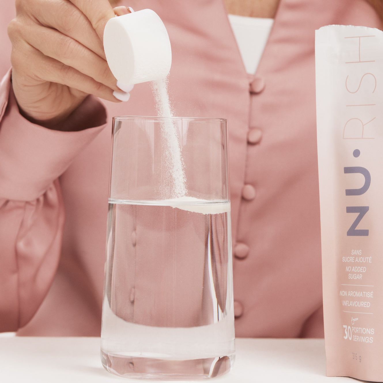 Main versant une mesure de poudre de collagène NU•RISH dans un verre d’eau, avec le sachet du produit visible à côté. Hand pouring a measure of NU•RISH collagen powder into a glass of water, with the product sachet visible next to it.