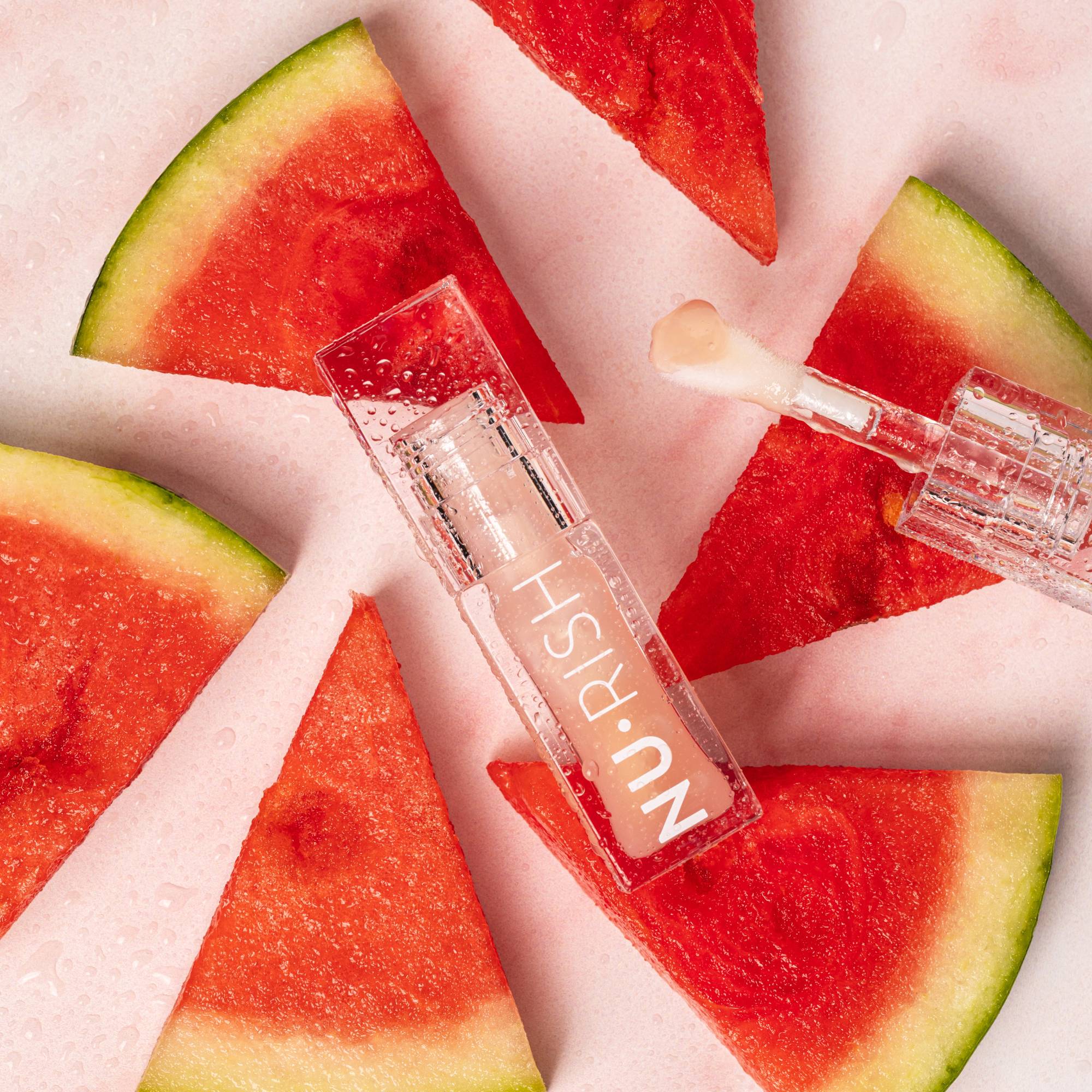 Huile à lèvres bioactive NU•RISH présentée avec son applicateur, entourée de tranches de melon d’eau fraîches sur un fond rosé. Un visuel estival qui évoque l’hydratation, la fraîcheur et la brillance des lèvres.