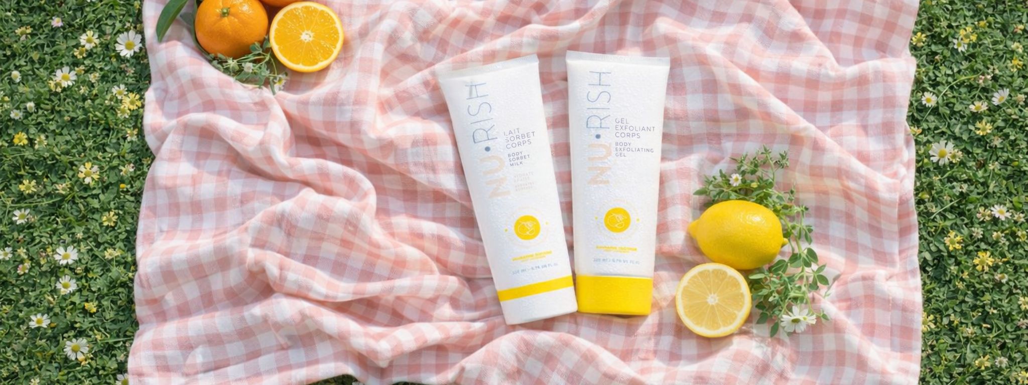 Photo en vue du dessus d’une mise en scène printanière : deux produits corps NU•RISH (lait sorbet hydratant et gel exfoliant) posés sur une nappe à carreaux rose pâle déposée sur de l’herbe fleurie. Autour, des citrons frais coupés et entiers, des fleurs blanches et de la verdure, dans une lumière naturelle douce évoquant la fraîcheur et la saison estivale.