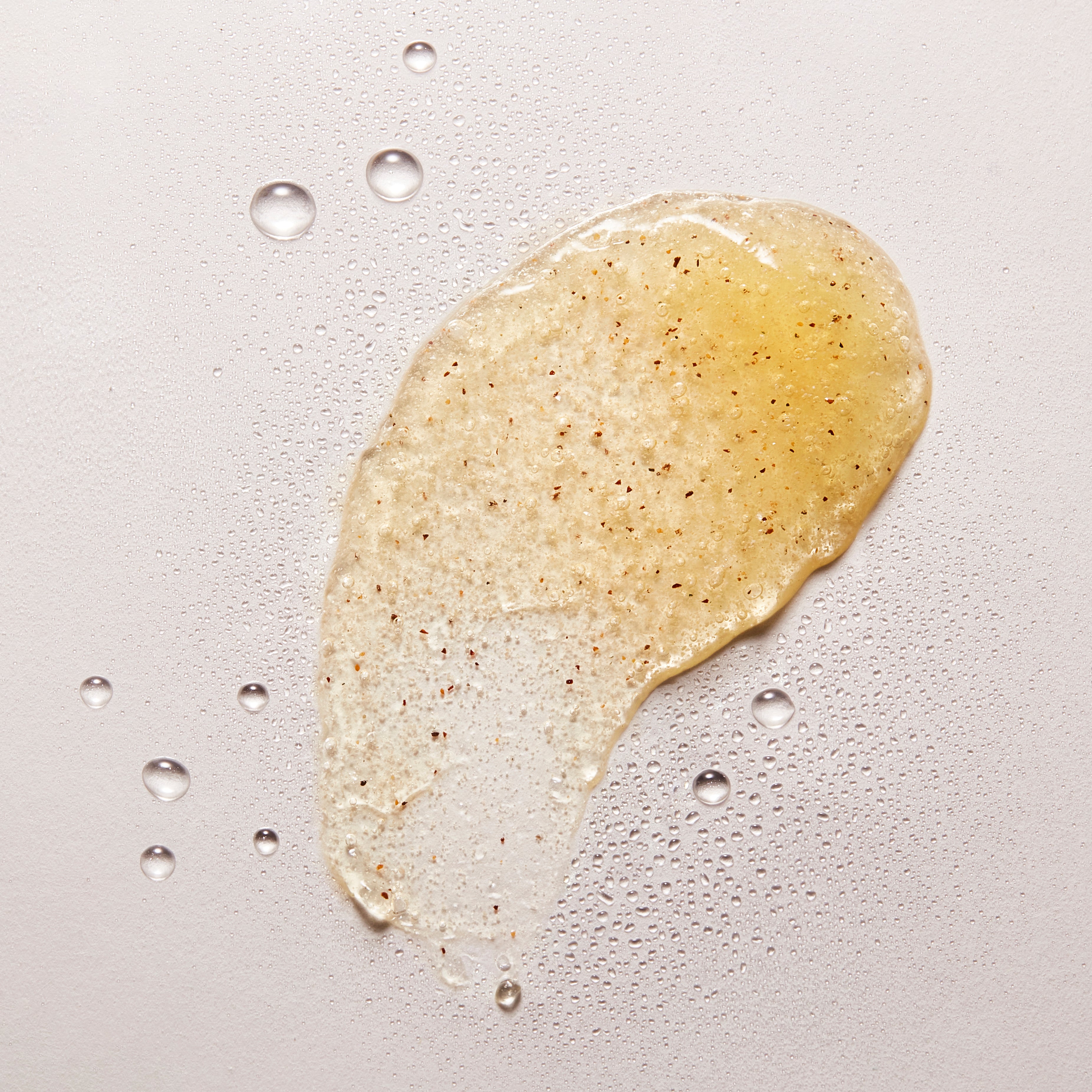 Texture de gel exfoliant NU•RISH appliquée sur fond clair, avec grains exfoliants visibles et gouttelettes d’eau, illustrant une exfoliation douce et hydratante. NU•RISH exfoliating gel texture applied to a light background, with visible exfoliating grains and water droplets, illustrating gentle, moisturizing exfoliation.